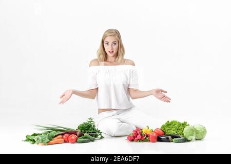 Une femme blonde frustrée se répand les mains dans les deux directions et beaucoup de légumes frais sur fond blanc Banque D'Images