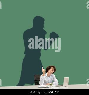 Un jeune homme caucasien rêve de l'avenir dans le grand sport pendant son travail au bureau. Ombre, silhouette de boxeur professionnel sur le mur. Devenir une légende. Inspiration, aspiration. CopySpace. Banque D'Images