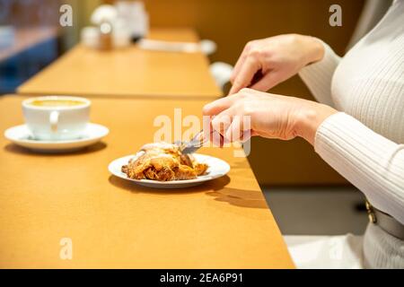 une femme coupe un croissant et boit du café à une table dans un café. pas de visage Banque D'Images