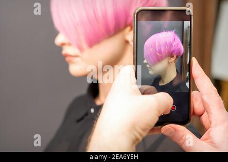 Le coiffeur prend des photos de la courte coiffure rose de une jeune femme sur le smartphone Banque D'Images