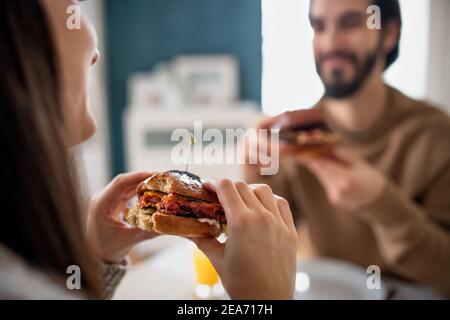 Un jeune couple méconnaissable adore manger des hamburgers à l'intérieur à la maison. Banque D'Images