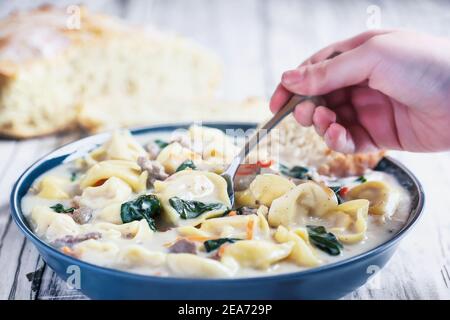 Main avec soupe Tortellini avec saucisse italienne, épinards et carottes. Servi avec du pain artisanal fait maison sur une table en bois blanc. Banque D'Images