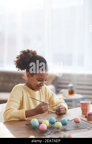 Portrait minimal de fille afro-américaine souriante à la main peignant des œufs de Pâques tout en appréciant la décoration de bricolage à la maison, espace de copie Banque D'Images