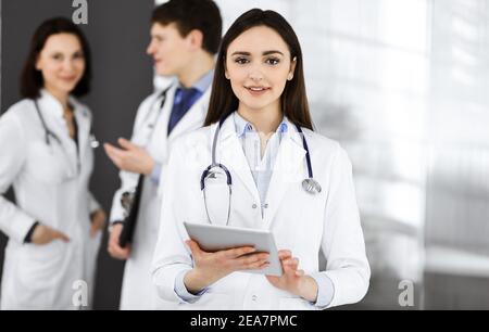 Une femme-médecin intelligente souriante tient une tablette dans ses mains, tandis qu'elle se tient debout avec ses collègues dans une clinique. Médecins Banque D'Images