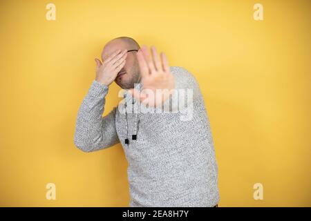 Jeune homme bald portant des lunettes sur fond jaune couvrant les yeux avec les mains et faisant un geste d'arrêt avec l'expression de la tristesse et de la peur. Embarrassé et ne Banque D'Images