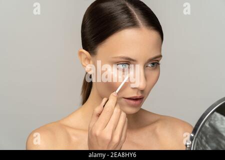 Femme avec maquillage naturel application de correcteur sur la peau fraîche impeccable, faisant maquillage regardant le miroir Banque D'Images