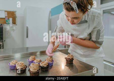 Gros plan de la confiserie féminine portant des gants roses pour préparer des cupcakes avec de la crème et des baies fraîches dans le studio de confiserie Banque D'Images