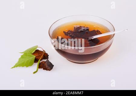 Morceaux de champignons chaga, bol à thé en verre décoré de feuilles de bouleau vert isolées sur fond blanc, gros plan. Inonotus obliquus. Banque D'Images