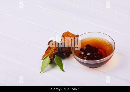 Morceaux de champignons chaga, bol à thé en verre décoré de feuilles de bouleau vert isolées sur fond blanc, gros plan. Inonotus obliquus. Banque D'Images