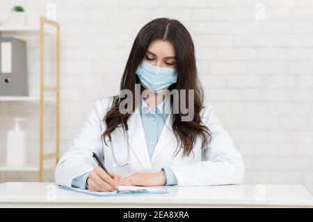 Jeune femme sérieuse médecin en manteau blanc et masque de protection écrit la prescription au patient de la clinique Banque D'Images