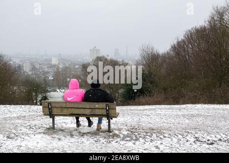 Un couple assis sur un banc de parc en bois qui regarde la vue de Londres depuis une colline enneigée du Parlement sur Hampstead Heath. La colline du Parlement est un point de vue populaire sur les gratte-ciel de la ville. Banque D'Images