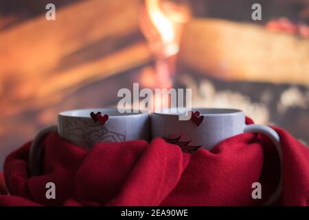 L'amour et la Saint-Valentin humeur romantique. Gros plan deux mugs à double cœur rouge sont attachés avec un foulard rouge. L'arrière-plan est le bois chaud dans la cheminée. Banque D'Images