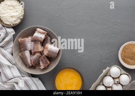 Dans un bol, des morceaux de merlu et des produits pour la cuisine frite, des œufs, de la farine et de la chapelure ont été présentés avec une serviette de cuisine sur fond gris Banque D'Images