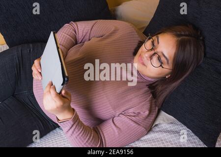 Une jeune femme latine s'est étirée sur son lit de chambre reposant sa tête sur un coussin bleu regardant la tablette Banque D'Images