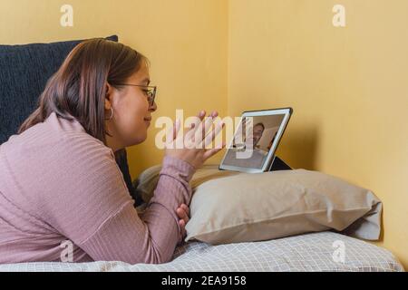 une jeune fille s'est étirée dans le lit en faisant un appel vidéo avec un gentleman, ils se saluent tous les deux par la caméra tablette Banque D'Images