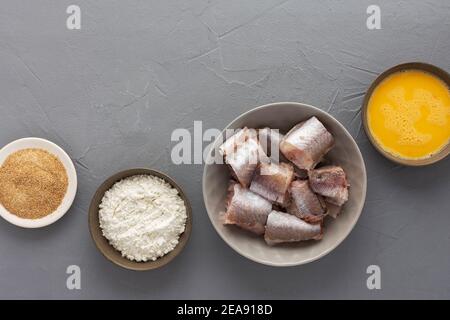 Dans un bol, des morceaux de merlu et des produits pour la cuisson frite, des œufs, de la farine et de la chapelure sur fond gris Banque D'Images