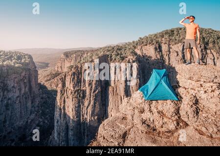 Homme randonneur ou grimpeur extrême se tient près de la tente de camping sur un point de vue de falaise au profond canyon Tazi en Turquie. Découvrez les merveilles de la nature et de la dange Banque D'Images