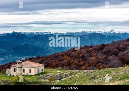 Italie Veneto Monte Baldo - la costruzione Baiti d'Ortigara ai limiti del fitto bosco acceso dal feuillage autunnale. Sullo sfondo le catène montuose ad ovest del lago di Garda|Italie Veneto Monte Baldo - le bâtiment de Baiti d'Ortigara au bord de la forêt dense éclairée par le feuillage d'automne. En arrière-plan, la montagne s'étend à l'ouest du lac de Garde. Un randonneur d'époque monte le dernier tronçon près du refuge de Chirico, derrière lui les profils des montagnes qui entourent le lac de Garde. Banque D'Images