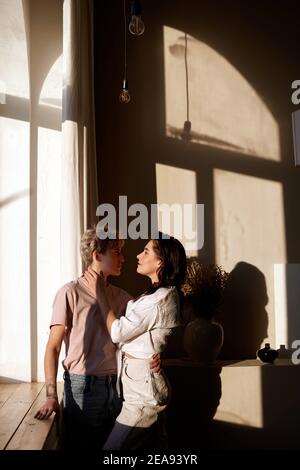 Vue latérale d'un couple amoureux qui se tient près de chez vous seuil de fenêtre dans la pièce éclairée par la lumière du soleil et regardant chacun autre Banque D'Images