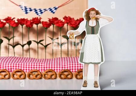 Photo de la vie encore, femme dans dirndl, avec bretzel à la main, mordant dans un bretzel, se dresse devant une décoration en bois, avec des bretzels, drapeaux bavarois, roses en plastique, couverture à carreaux sur le sol Banque D'Images