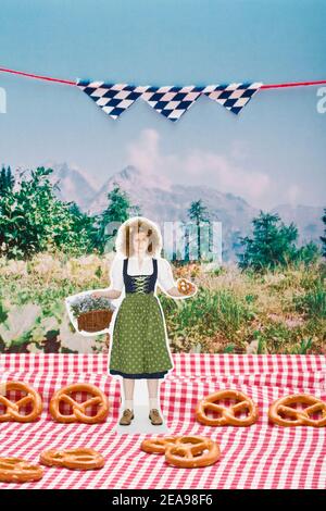 Femme dans dirndl, avec bretzel et panier de fleurs dans sa main, se tient sur une nappe à carreaux, devant une photo de prairie verte et de montagnes, les drapeaux bleus et blancs pendent dans le fond, bretzels sont sur le sol, encore la vie Banque D'Images