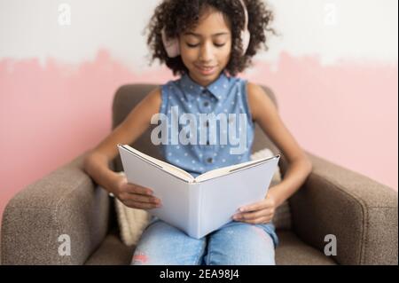 Smart biracial pré-école fille de passer du temps de loisirs avec un lecture de livres assis à un fauteuil à la maison Banque D'Images