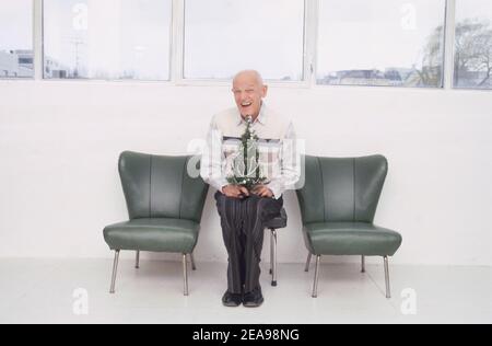 Le vieil homme est assis avec un petit arbre de Noël sur une chaise entre deux fauteuils Banque D'Images