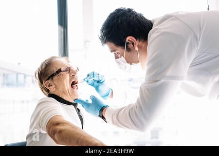 Vue latérale du médic mâle dans le masque de protection prenant COVID 19 tests réalisés chez une patiente senior à domicile Banque D'Images