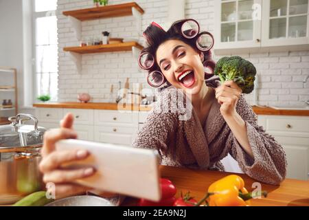 Bonne jeune femme en curlers prenant un selfie drôle avec le brocoli chef dans la cuisine Banque D'Images