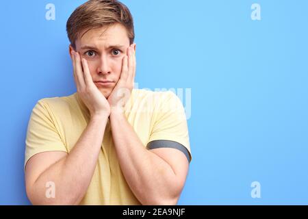 triste homme tenant les joues et regardant la caméra avec l'expression mélancholique. isolé fond bleu. portrait de jeunes hommes caucasiens dans un t-shirt décontracté Banque D'Images