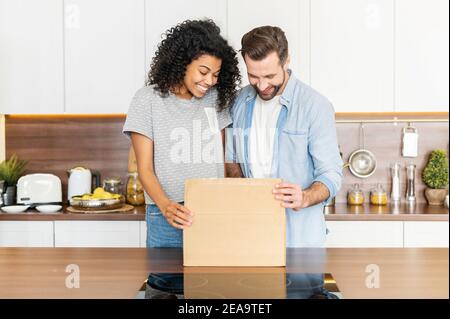 Heureux couple interracial souriant debout près de la table dans la cuisine moderne et ouvrant une boîte en carton, jeune famille millénaire excitée de recevoir Banque D'Images