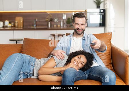 Couple interracial se reposant sur le canapé, regardant un film dans l'appartement studio, souriant une jeune femme afro-américaine surjoyeuse couché sur elle Banque D'Images