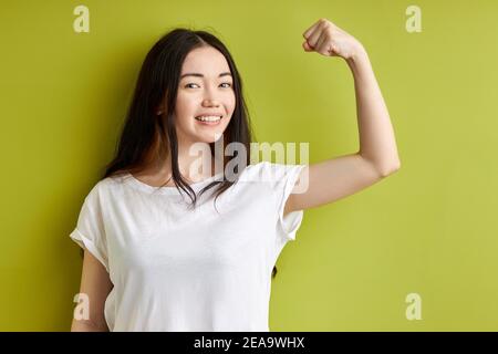 la jeune femme lève le bras et montre biceps a l'expression heureuse démontre ses réalisations après son entraînement dans la salle de gym habillée décontractée isolée sur l'ba vert Banque D'Images