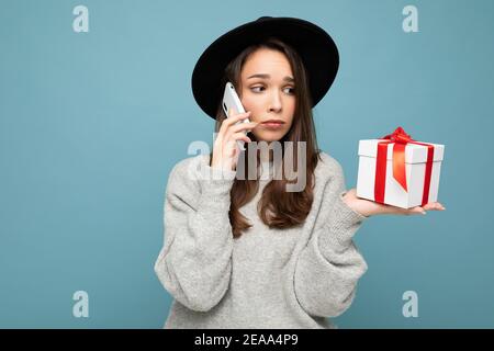 Photo de la jeune femme brunette bouleversée belle isolée sur bleu mur d'arrière-plan portant un chapeau noir et un chandail gris tenant un cadeau box parlant sur mobile Banque D'Images