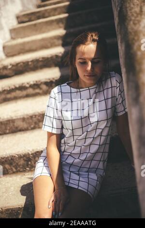 Jeune femme dans l'ombre assise sur des escaliers en béton à l'extérieur à la journée ensoleillée. Concept de style de vie urbain Banque D'Images