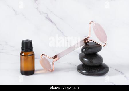 Rouleau de jade massage facial aux quartes roses avec huile cosmétique et pierres de basalte Zen noires sur fond de marbre blanc. Soins du visage. Banque D'Images