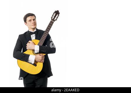 Beau jeune homme en costume classique noir avec noeud papillon posant avec guitare classique en studio sur fond blanc. Banque D'Images