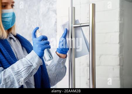 Femme de ménage portrait en caoutchouc bleu gants nettoyer le bouton de porte à l'aide d'un chiffon. Nettoyage de la poignée de porte avant à l'aide d'un aérosol antibactérien. Nouvelle normale Banque D'Images