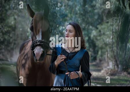 Fille menant un cheval à travers les arbres tout en portant un robe bleue de style médiéval dans le doux bleuâtre matin ensoleillé léger Banque D'Images