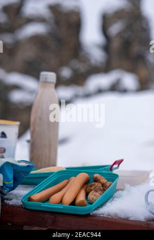 Pique-nique en hiver, les saucisses sont posées sur une table dans une boîte de Tupperware. Il y a un thermos à côté. Banque D'Images