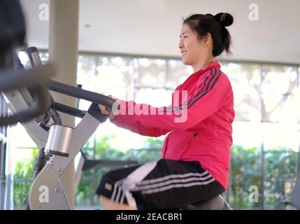 Une jeune femme asiatique attirante fait de l'exercice dans une salle de sport, à vélo d'appartement pour se réchauffer avant de se déplacer vers d'autres appareils de gym. Banque D'Images