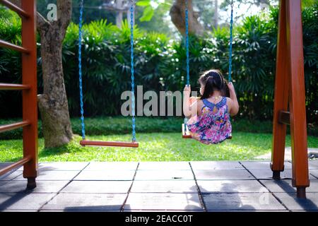 La vue arrière d'une jeune fille asiatique solitaire jouant sur une balançoire par elle-même dans une aire de jeux vide par une journée ensoleillée. Banque D'Images