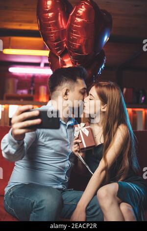 Couple caucasien embrassant sur un canapé rouge tenant des ballons d'air et faire un selfie avec le téléphone Banque D'Images