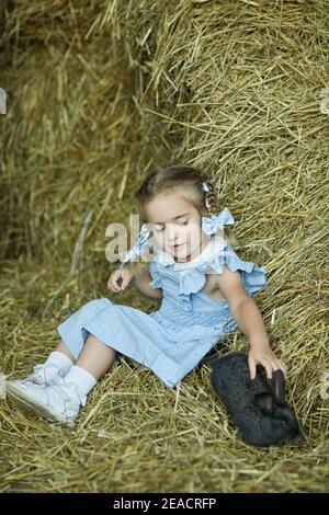 Petite jolie fille est assise dans le foin avec un petit lapin. La fille nourrit son animal de compagnie préféré. Été dans un village. Bonne enfance Banque D'Images