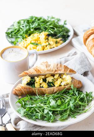 Délicieux sandwich croissant avec œufs d'épinards brouillés servi avec de l'arugula sur l'assiette et une tasse de café, fond en béton léger. Mise au point sélective. Banque D'Images