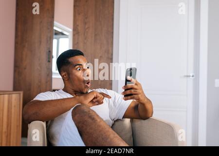 Un homme afro-américain stupéfait assis dans un fauteuil confortable et utilisant un téléphone portable. Banque D'Images