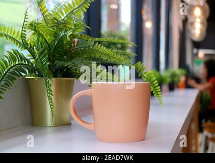 Tasse à café orange pastel avec fougères en pot sur une longue table avec des personnes floues assis à distance Banque D'Images