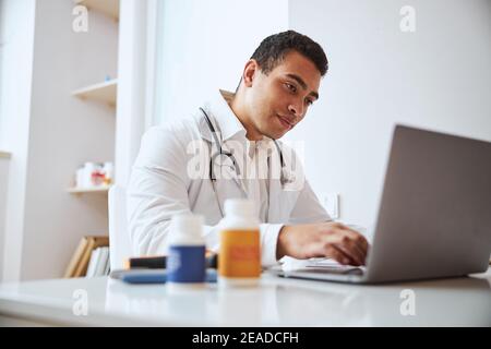 homme charmant et confiant thérapeute en robe blanche avec stéthoscope assis au bureau de travail dans l'armoire Banque D'Images