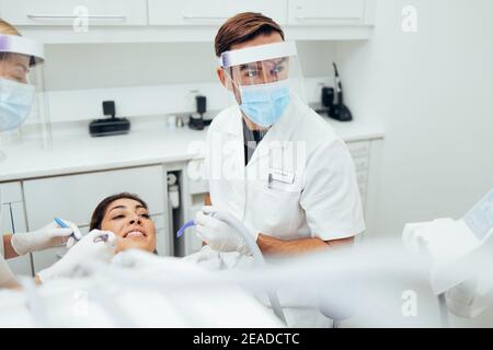 Dentiste traitant une patiente à la clinique dentaire. Un médecin de sexe masculin portant un masque et un écran facial pendant le traitement dentaire d'une femme. Banque D'Images