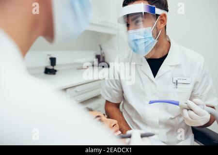 Les médecins portant des écrans faciaux discutant entre le traitement dentaire d'une patiente. Dentiste masculin tenant le tube d'aspiration discutant avec son assistant Banque D'Images